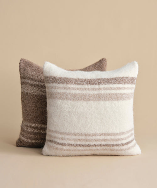 Two square throw pillows rest on a beige surface: one is a cream-striped Sheridan Pillow, and the other is solid brown with a fuzzy alpaca blend texture.