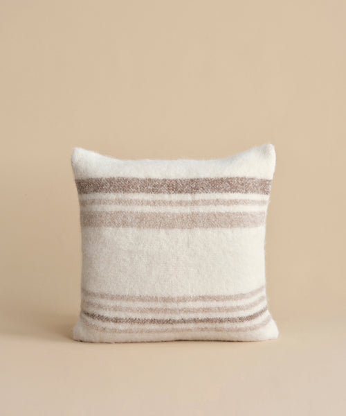 The Sheridan Pillow, a square cushion featuring horizontal beige and light brown stripes on white alpaca blend yarn, is shown upright against a plain beige background.