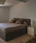 A minimalist bedroom featuring a bed with a dark brown upholstered headboard and skirt, dressed in taupe bedding and two matching striped pillows, set against a neutral gray wall.