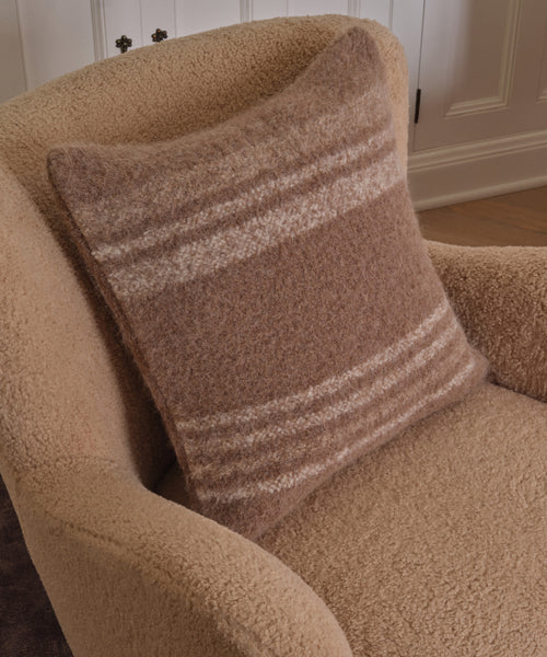 A textured beige armchair with the Sheridan Pillow, an alpaca blend cushion featuring horizontal brown and cream stripes, set in front of a white paneled wall.