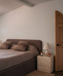 A bedroom scene featuring a bed with brown upholstered headboard and matching brown bedding, styled with two brown striped pillows. A light wood nightstand with a white table lamp and a small candle sits beside the bed against a warm off-white wall.