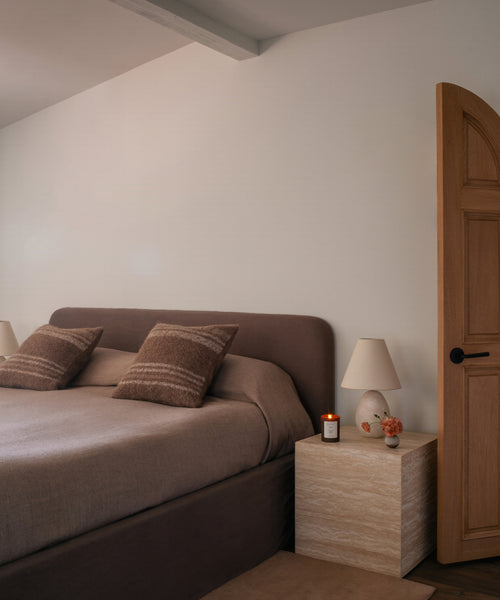 A bedroom scene featuring a bed with brown upholstered headboard and matching brown bedding, styled with two brown striped pillows. A light wood nightstand with a white table lamp and a small candle sits beside the bed against a warm off-white wall.