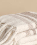 A close-up of the Sheridan Throw, a soft folded blanket crafted from luxurious alpaca blend yarn, featuring white and light beige stripes against a plain beige background.