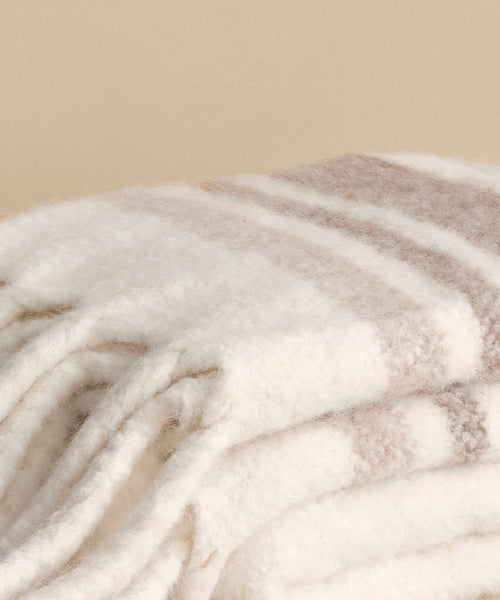 A close-up of the Sheridan Throw, a soft folded blanket crafted from luxurious alpaca blend yarn, featuring white and light beige stripes against a plain beige background.