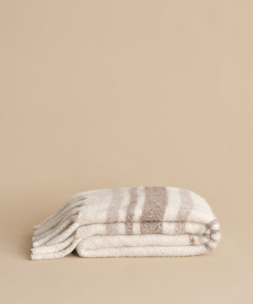 The Sheridan Throw is a folded, cozy blanket crafted from an alpaca blend yarn with light beige and cream horizontal stripes and fringed edges, displayed on a neutral beige background.