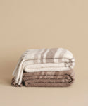 Two Sheridan Throws stacked on a beige background: the top is striped cream and brown, the bottom solid light brown—both made with soft, warm alpaca blend yarn, perfect for cozy accents in any home.