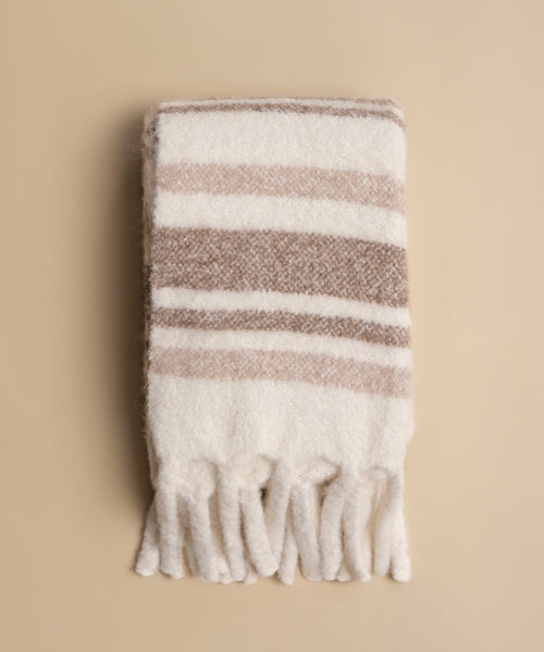 The Sheridan Throw, a soft alpaca blend with beige and white stripes and fringe tassels, is folded and displayed on a neutral beige background.