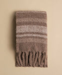 The Sheridan Throw, crafted from an alpaca blend, is a folded fuzzy brown scarf with light beige stripes and thick twisted fringes on the ends, shown on a plain beige background.