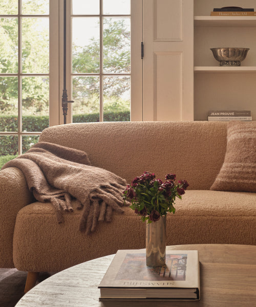 A cozy living room features a beige textured sofa adorned with a Sheridan Throw, a matching pillow, a wooden coffee table topped with books and wilted flowers, and large windows filling the space with natural light.