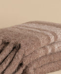 A close-up of the Sheridan Throw, a soft, brown woolen blanket with subtle light stripes, crafted from alpaca blend yarn and shown folded on a neutral beige surface.