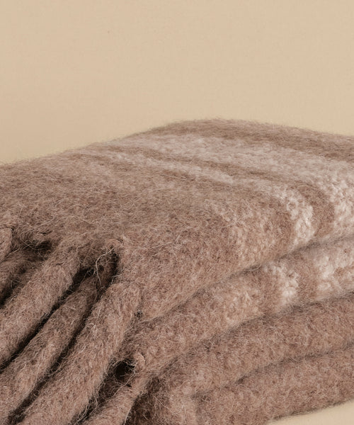 A close-up of the Sheridan Throw, a soft, brown woolen blanket with subtle light stripes, crafted from alpaca blend yarn and shown folded on a neutral beige surface.