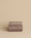 The Sheridan Throw, crafted from a soft alpaca blend yarn in brown and beige stripes, is elegantly folded and displayed against a plain beige background.