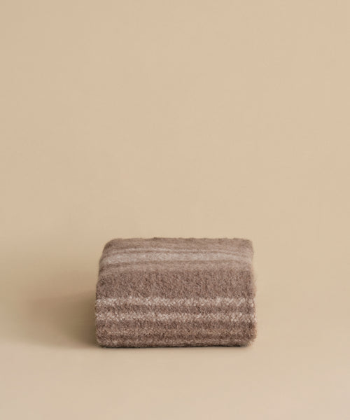 The Sheridan Throw, crafted from a soft alpaca blend yarn in brown and beige stripes, is elegantly folded and displayed against a plain beige background.