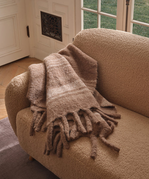 A soft, textured beige armchair features the Sheridan Throw—a thick, brown fringed striped blanket—draped over one arm as sunlight pours through glass doors, brightening the cozy living space.