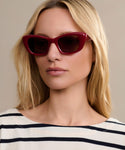 A woman with long blonde hair wears Sierra Sunglasses—retro cat eye shades offering 100% UV protection—and a white top with black stripes, looking to the side against a beige background.