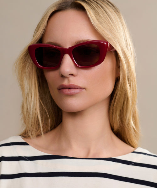 A woman with long blonde hair wears Sierra Sunglasses—retro cat eye shades offering 100% UV protection—and a white top with black stripes, looking to the side against a beige background.