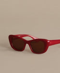 The Sierra Sunglasses by Garret Leight California Optical feature red retro cat-eye frames, dark lenses with 100% UV protection, set stylishly on a neutral beige surface and angled slightly to the right.