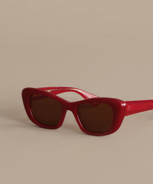 The Sierra Sunglasses by Garret Leight California Optical feature red retro cat-eye frames, dark lenses with 100% UV protection, set stylishly on a neutral beige surface and angled slightly to the right.