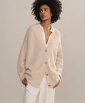 A woman with curly hair stands against a neutral background, wearing the Silk Sloane Shirt in ivory under a loose, beige cardigan with white pants. She looks to the side with one hand in her pocket.