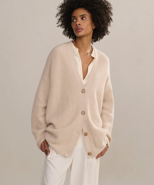 A woman with curly hair stands against a neutral background, wearing the Silk Sloane Shirt in ivory under a loose, beige cardigan with white pants. She looks to the side with one hand in her pocket.