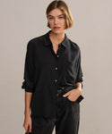 A woman with short, light brown hair wears the Silk Sloane Shirt—relaxed fit, black, with rolled sleeves—and matching black pants. She stands against a plain light background, confidently looking at the camera with one hand in her pocket.