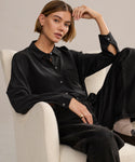 A woman with short brown hair sits in a cream chair, wearing the relaxed fit Silk Sloane Shirt and black jeans. One arm rests on the chair as she looks calmly at the camera.