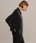 A person with short light brown hair stands sideways, looking down, wearing the relaxed fit black Silk Sloane Shirt and black jeans against a plain beige background.
