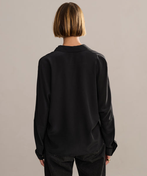A person with short, straight blonde hair, seen from behind, wears the Silk Sloane Shirt in black and dark pants while standing against a plain beige background.