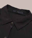 Close-up of the Silk Sloane Shirt in black, featuring smooth silk fabric, a relaxed fit, button front, and a visible black label with subtle text inside the collar, shown on a beige background.