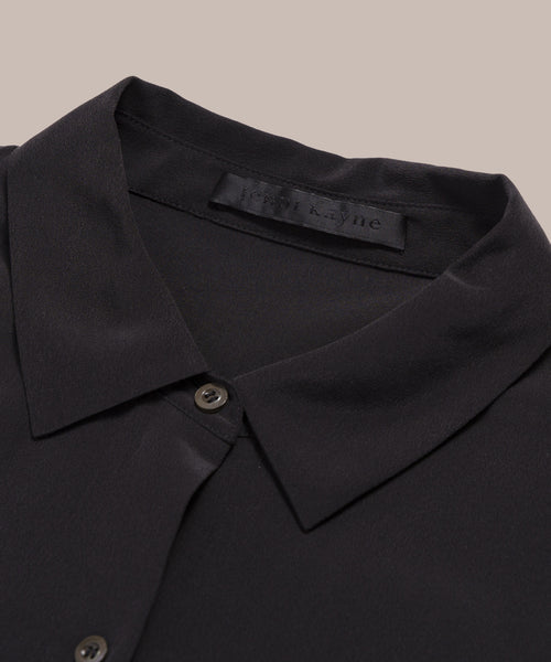 Close-up of the Silk Sloane Shirt in black, featuring smooth silk fabric, a relaxed fit, button front, and a visible black label with subtle text inside the collar, shown on a beige background.