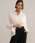 A woman with short brown hair wears the loose, ivory Silk Sloane Shirt tucked into high-waisted black pants with a black belt. She stands against a plain light background, gazing into the camera with a relaxed expression.