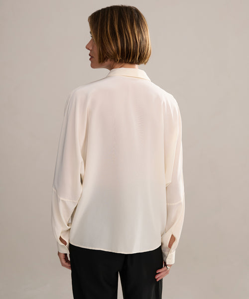 Wearing the Silk Sloane Shirt in ivory and black pants, a person with short light brown hair stands facing away from the camera against a plain light background.