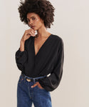 A woman with natural curly hair wears the Silk Sydney Top, a black V-neck long sleeve shirt, paired with blue jeans. She poses confidently—one hand near her face, the other in her pocket—against a plain light background.