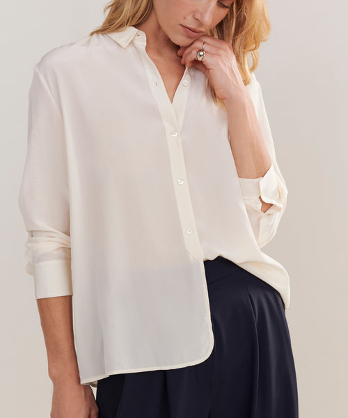 A woman wears the Silk Violette Shirt, a relaxed button-up with long sleeves in white, paired with dark trousers. One hand touches her hair while the other rests at her side.