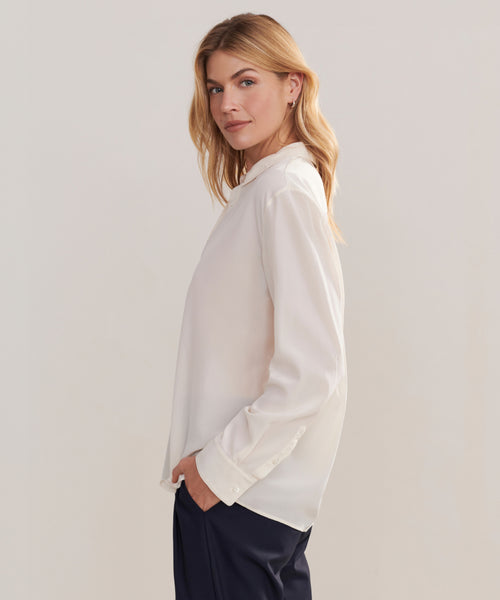 A woman with long blonde hair models the Silk Violette Shirt and dark pants, standing sideways with one hand in her pocket and looking over her shoulder at the camera against a plain background.