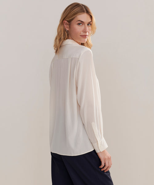 A woman with shoulder-length blonde hair wears the Silk Violette Shirt, a relaxed white button-up blouse, paired with dark pants as she stands sideways and looks over her shoulder against a plain background.