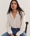 A woman with long dark hair sits on a chair, wearing the Silk Violette Shirt and blue jeans. She gazes calmly at the camera, set against a plain background.