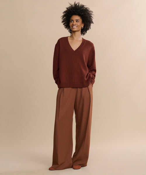 A woman with curly hair smiles in the Simone V-Neck, a loose dark brown cotton merino sweater, paired with wide-legged light brown trousers, standing against a plain beige background.
