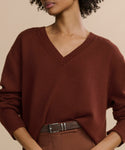 A person wears the Simone V-Neck, a loose-fitting cotton merino sweater in rust, tucked into high-waisted brown pants with a matching belt against a neutral background—effortlessly luxurious for any occasion.
