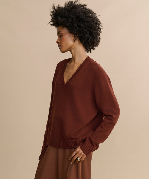 Woman with curly hair wears the Simone V-Neck in rust, paired with a brown skirt, standing sideways against a beige background and looking forward with a relaxed expression—a luxurious layer for any occasion.