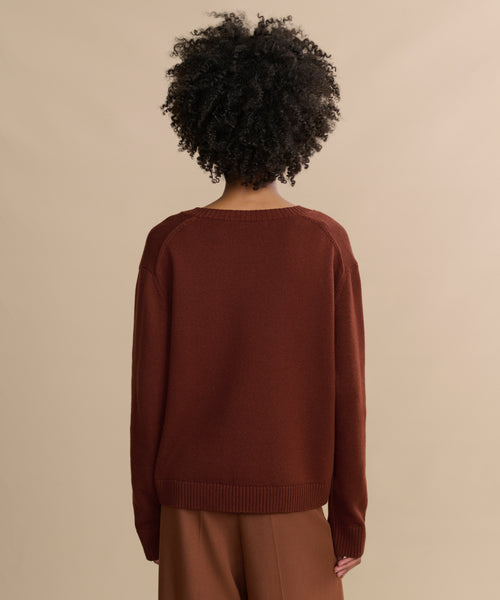 Seen from behind, a person with curly hair wears the Simone V-Neck sweater and brown pants—a loose-fitting luxury layer styled against a plain beige background.