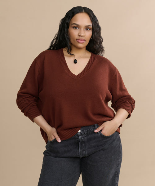 A woman with long, wavy hair poses confidently against a beige background wearing the rust-colored Simone V-Neck sweater, dark jeans, and a black pendant necklace. Her hands are in her pockets.