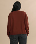 Seen from the back, a person with long, wavy dark hair wears the Simone V-Neck in rust—a soft cotton merino blend sweater—with dark gray jeans, standing against a plain beige background.