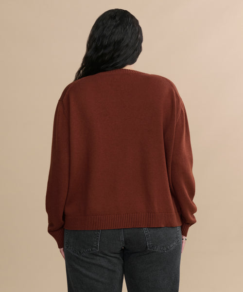 Seen from the back, a person with long, wavy dark hair wears the Simone V-Neck in rust—a soft cotton merino blend sweater—with dark gray jeans, standing against a plain beige background.