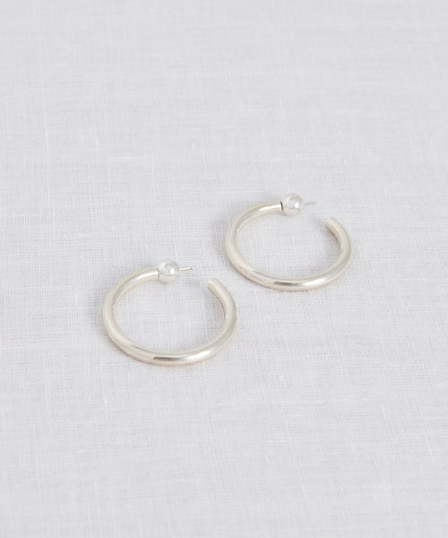 The Small Clara Hoops, crafted in sterling silver with post backings, are displayed on a light, textured fabric surface.