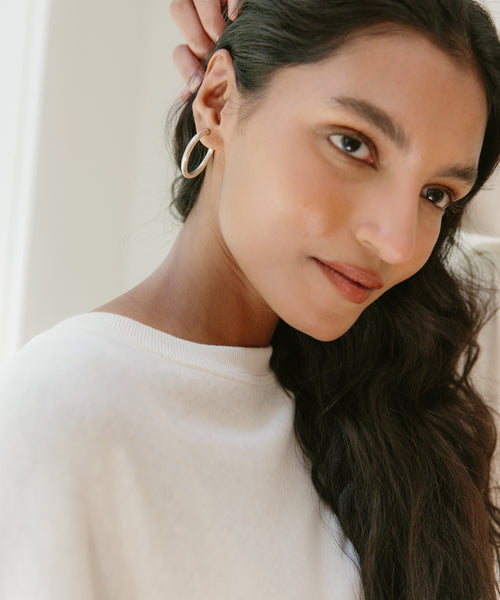 A woman with long dark hair, wearing a white top and Sophie Buhai Small Clara Hoops in sterling silver, touches her hair near her ear and gazes softly to the side with a natural expression.