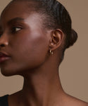 A close-up of a woman’s profile against a beige background highlights her smooth skin, braided bun, calm expression, and Jenni Kayne Small Eloise Hoops—timeless 14k gold heirloom jewelry.