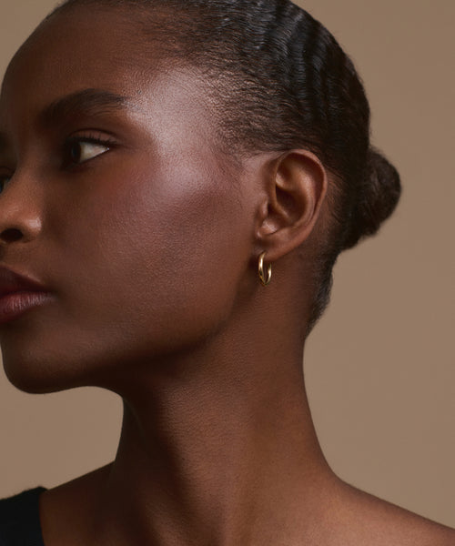 A close-up of a woman’s profile against a beige background highlights her smooth skin, braided bun, calm expression, and Jenni Kayne Small Eloise Hoops—timeless 14k gold heirloom jewelry.