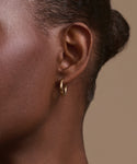 Close-up of a person’s ear adorned with Jenni Kayne Small Eloise Hoops in 14k gold. Hair is pulled back, highlighting smooth skin and the elegant, timeless touch of this heirloom jewelry against a plain beige background.