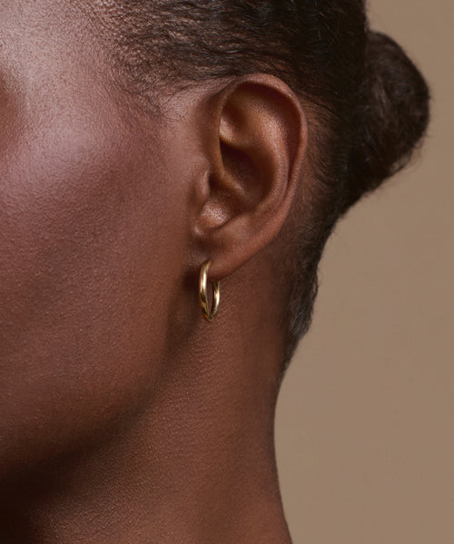 Close-up of a person’s ear adorned with Jenni Kayne Small Eloise Hoops in 14k gold. Hair is pulled back, highlighting smooth skin and the elegant, timeless touch of this heirloom jewelry against a plain beige background.